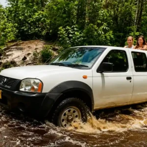 A white Nissan pickup truck driving through a muddy river in a dense green jungle, with three women riding in the open truck bed.