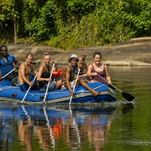 Een groep toeristen in een blauwe opblaasbare raft peddelt op een rustige rivier in Suriname, met dichte jungle op de achtergrond.