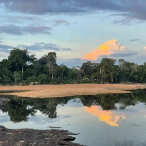 Een rustig rivierenlandschap in Suriname met een zandbank, dicht regenwoud en een weerspiegeling van oranje verlichte wolken in het water.